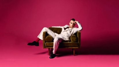 Portrait of Robbie Williams in a white suit on a velvet chair against a pink background at Moco Museum London